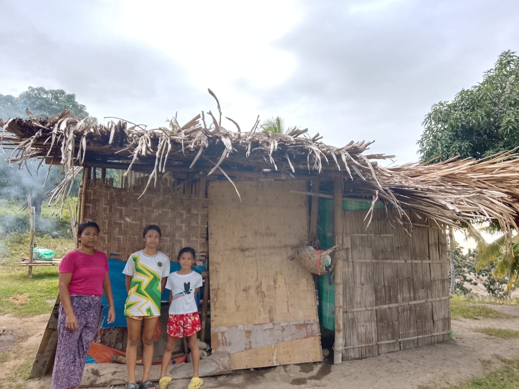 Loyren standing with her two daughters in front of a small bamboo and palm-leaf hut in the Philippines